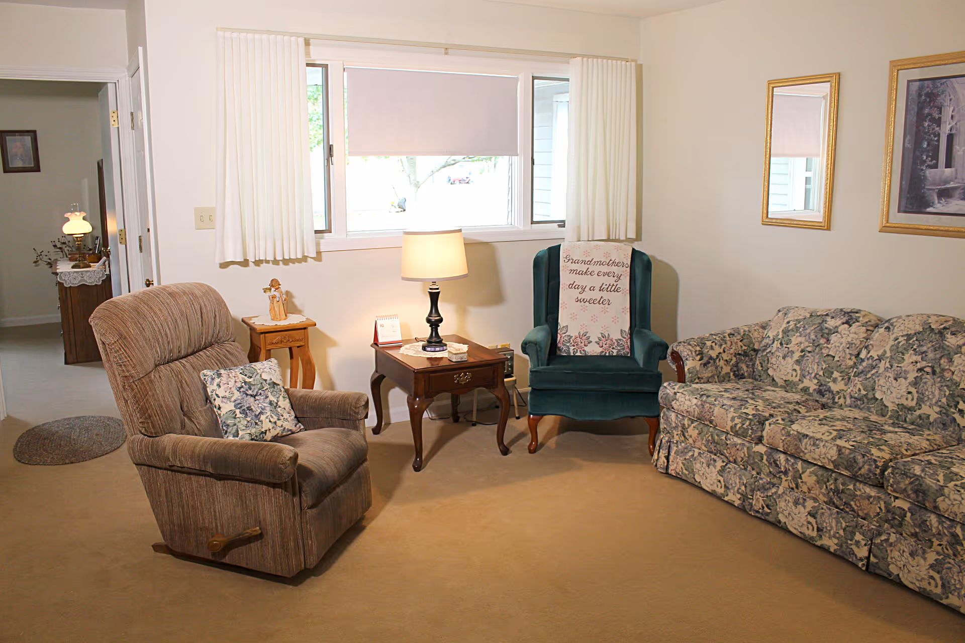 Living room with a brown recliner, wooden side tables and lamp, a green wingback chair, and a floral-patterned sofa beneath a window.
