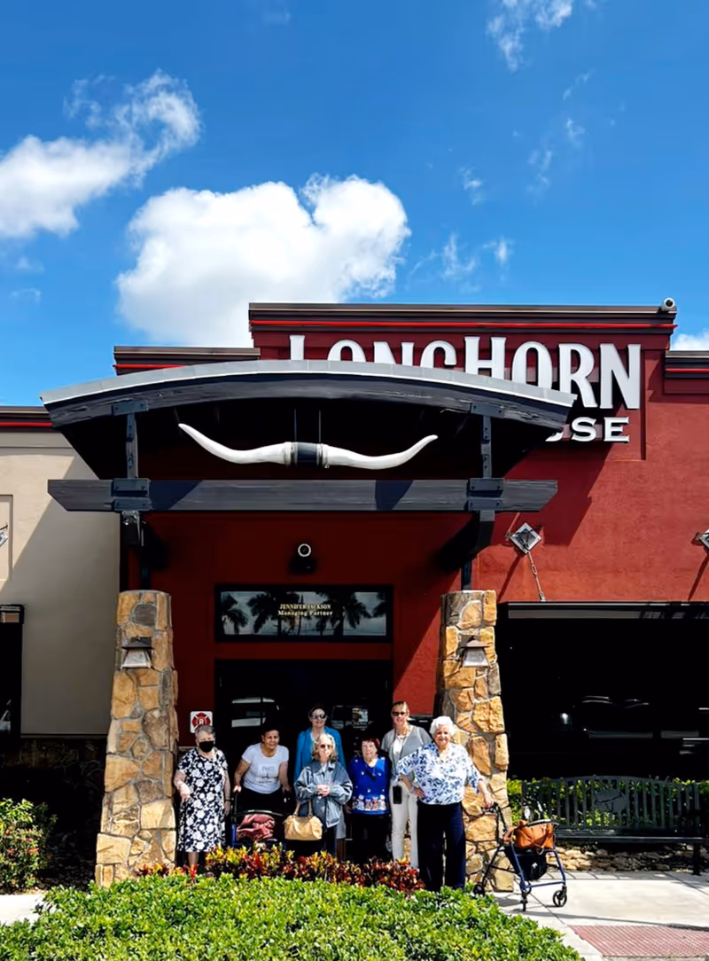 A group of seven people standing in front of the entrance to a LongHorn Steakhouse restaurant with a large longhorn decoration above the doorway. The building has stone pillars and a red facade under a blue sky with some clouds.