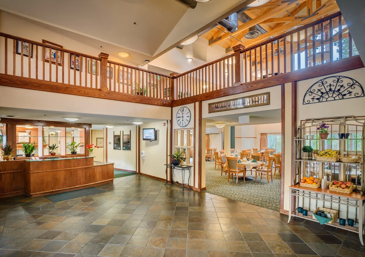 Interior view of Eskaton Village Grass Valley showing a spacious lobby area with a wooden reception desk on the left, a large clock on the wall, and an open dining room with tables and chairs set for meals on the right. The ceiling features wooden beams and a ceiling fan, with a second-floor balcony railing visible above.