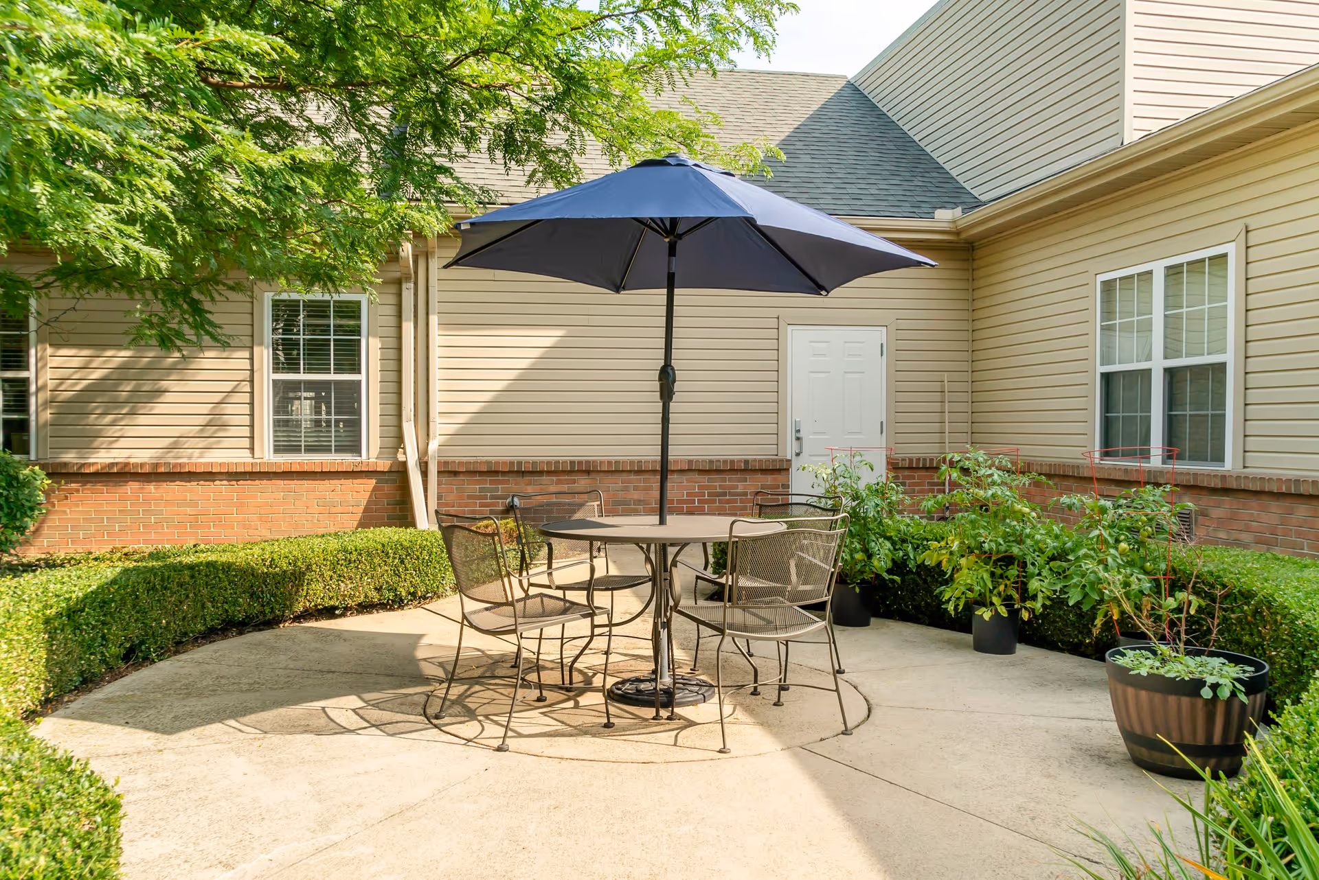 Outdoor patio area with a round metal table and four matching chairs under a large blue umbrella. The patio is surrounded by neatly trimmed bushes and potted plants, with beige siding and brick exterior walls of a building in the background.