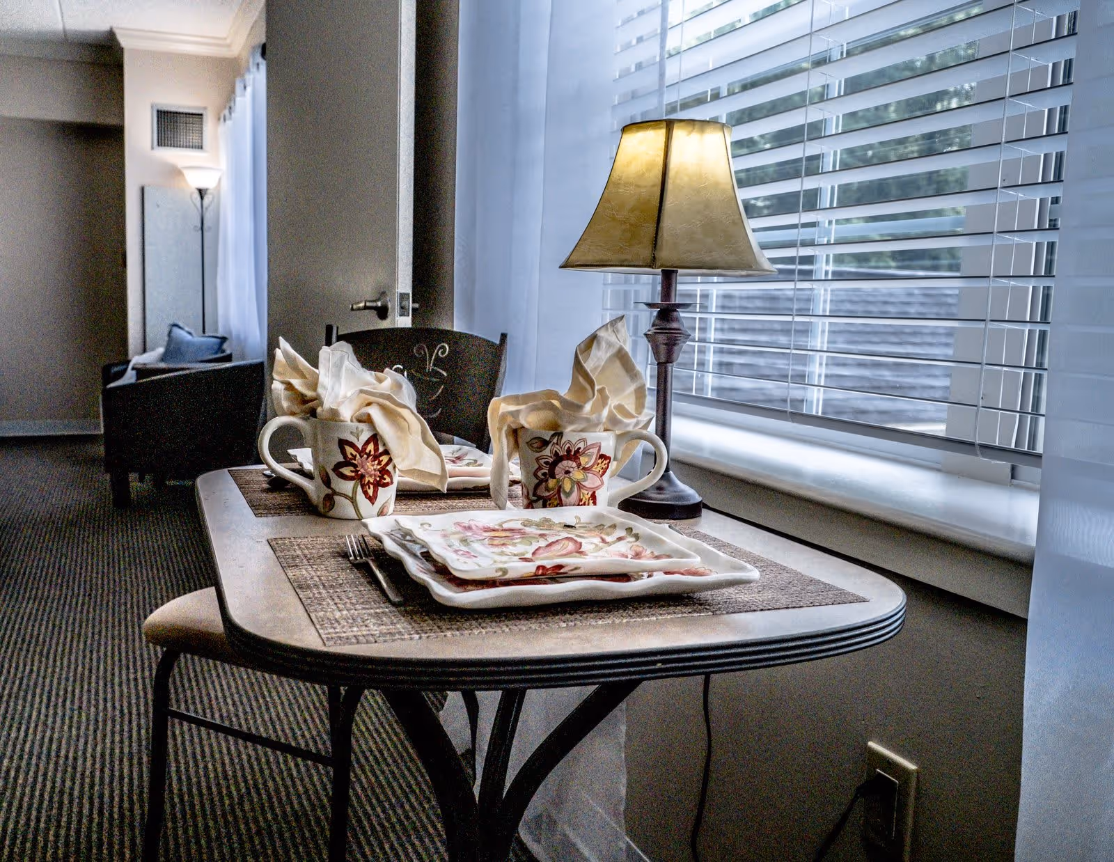 A small dining table set for two with floral-patterned plates and mugs, cloth napkins folded inside the mugs, and a table lamp beside the window with blinds partially open. In the background, there is a chair and a floor lamp in a softly lit room.
