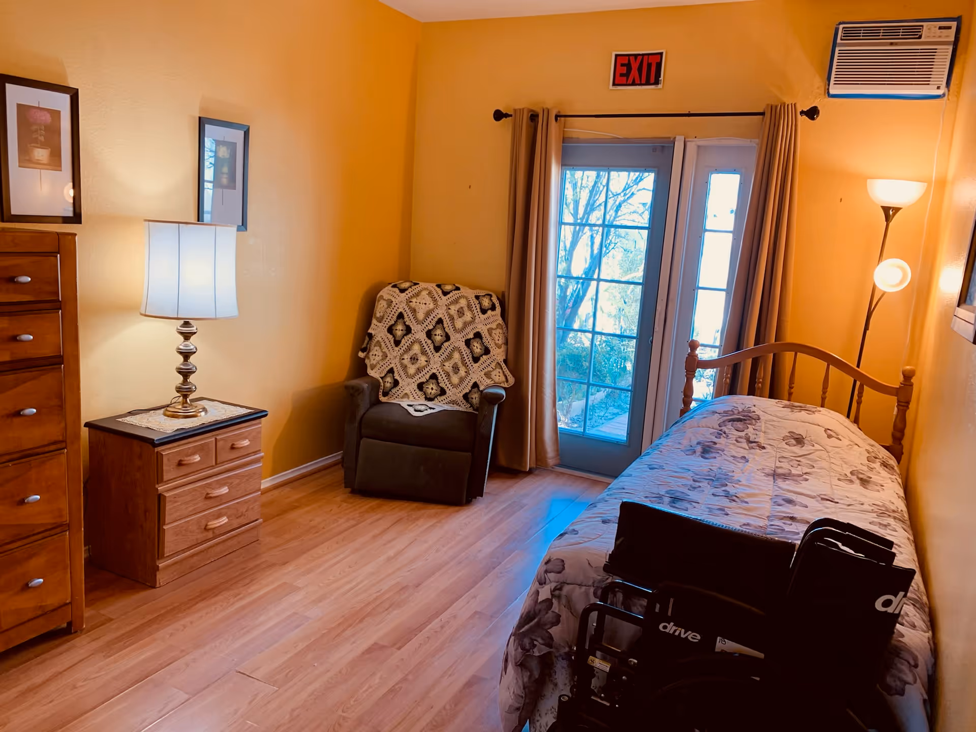 A cozy bedroom with yellow walls and wooden flooring. The room features a single bed with floral bedding, a wooden dresser, a nightstand with a lamp, a cushioned armchair with a crocheted blanket, and a wheelchair in front of the bed. There is a glass door with curtains leading outside, an exit sign above the door, and an air conditioning unit mounted on the wall.