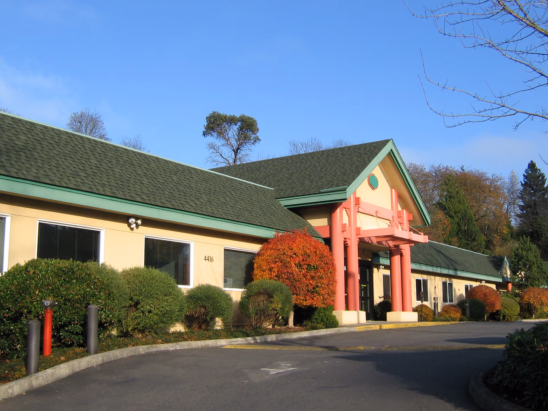 Exterior view of a single-story building with a green roof and beige walls, featuring a prominent entrance with red columns. The building is surrounded by neatly trimmed bushes and trees with autumn foliage under a clear blue sky.