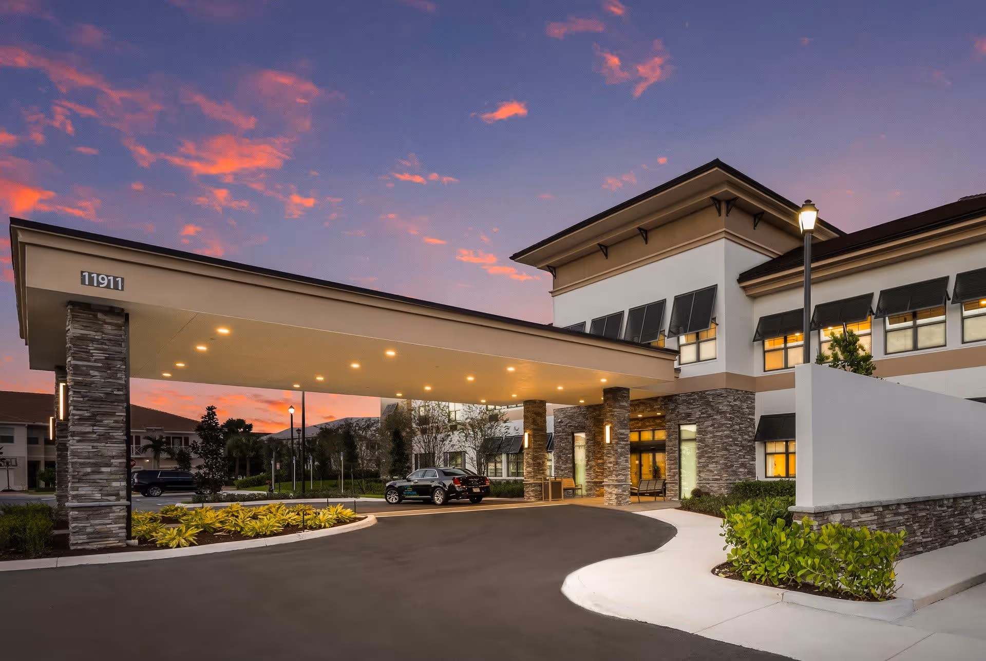 Front entrance and porte-cochere of a modern senior living building at dusk, with an illuminated canopy, stone columns, landscaping, and a parked car.