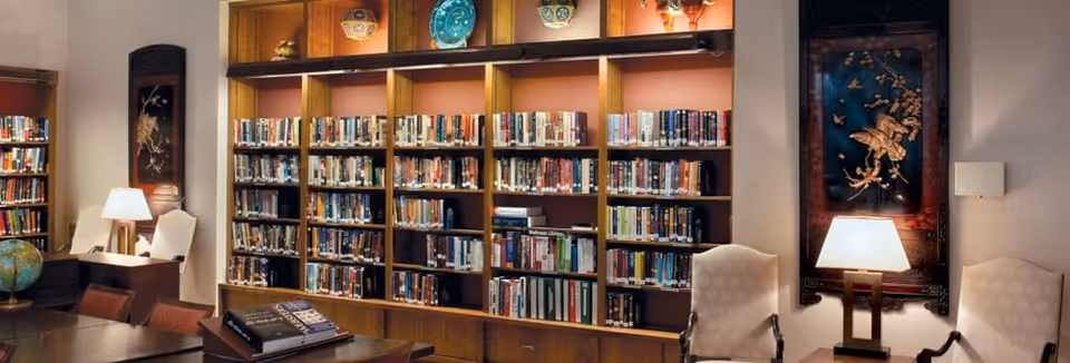 A cozy library room with wooden bookshelves filled with books, two white cushioned chairs, a table with a globe and books, and two table lamps providing warm lighting. Decorative plates and framed artwork are displayed on the walls.