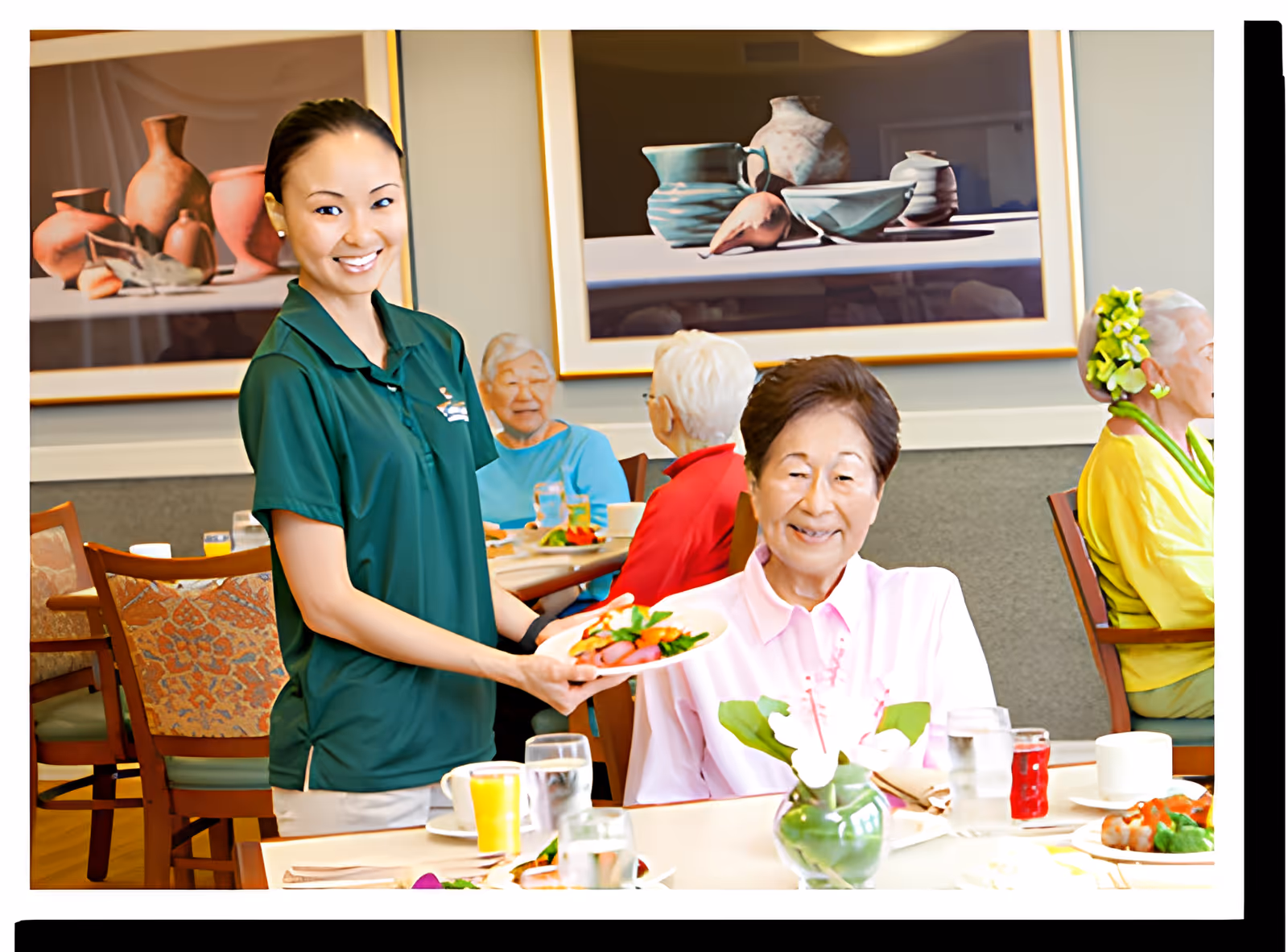A smiling staff member in a green polo shirt serves a plate of food to an elderly woman seated at a dining table in a senior living facility. Other elderly residents are seated at tables in the background, with framed artwork on the walls.