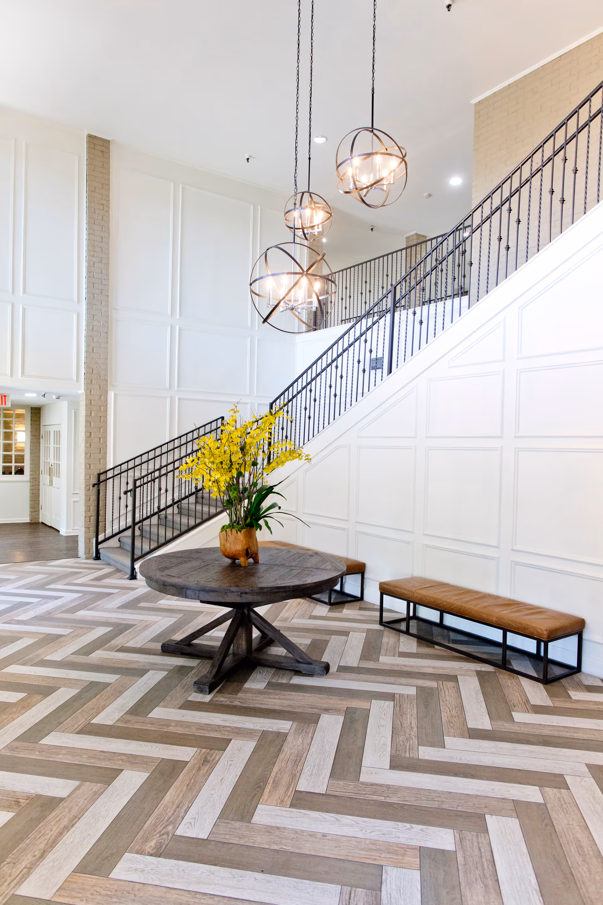 Bright and spacious interior area with a wooden round table holding a vase of yellow flowers in the center. There are two brown cushioned benches with black metal frames along the white paneled wall. A staircase with black metal railing ascends along the wall, and three modern spherical pendant lights hang from the ceiling. The floor features a herringbone pattern in various shades of wood.