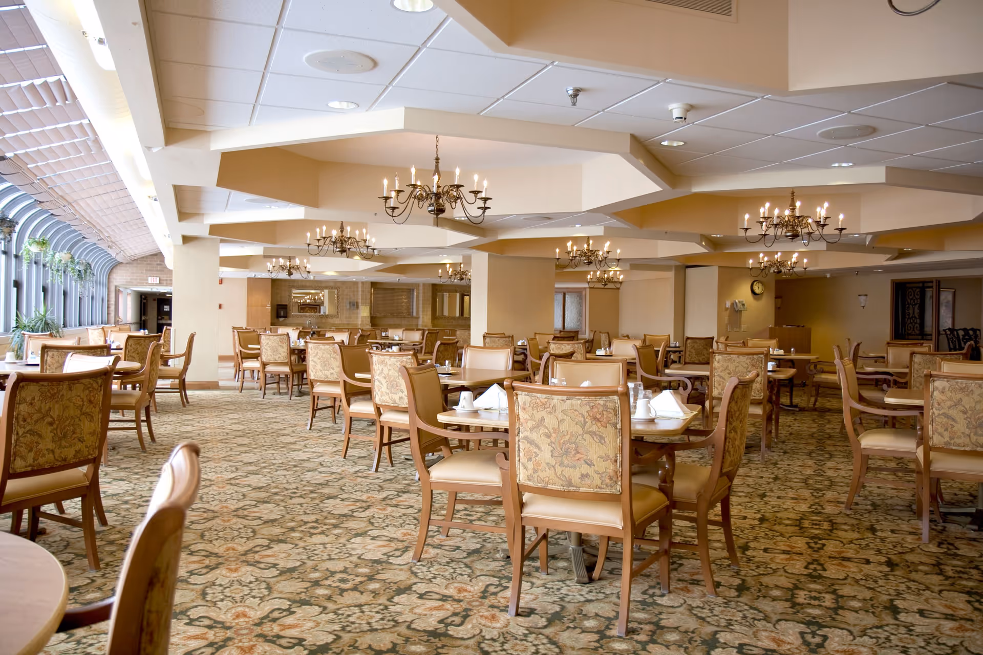 Spacious dining room with multiple round tables and cushioned chairs arranged neatly. The room features patterned carpet flooring, chandeliers hanging from the ceiling, and large windows along one side allowing natural light to enter. Tables are set with white napkins and cups, creating an inviting atmosphere.