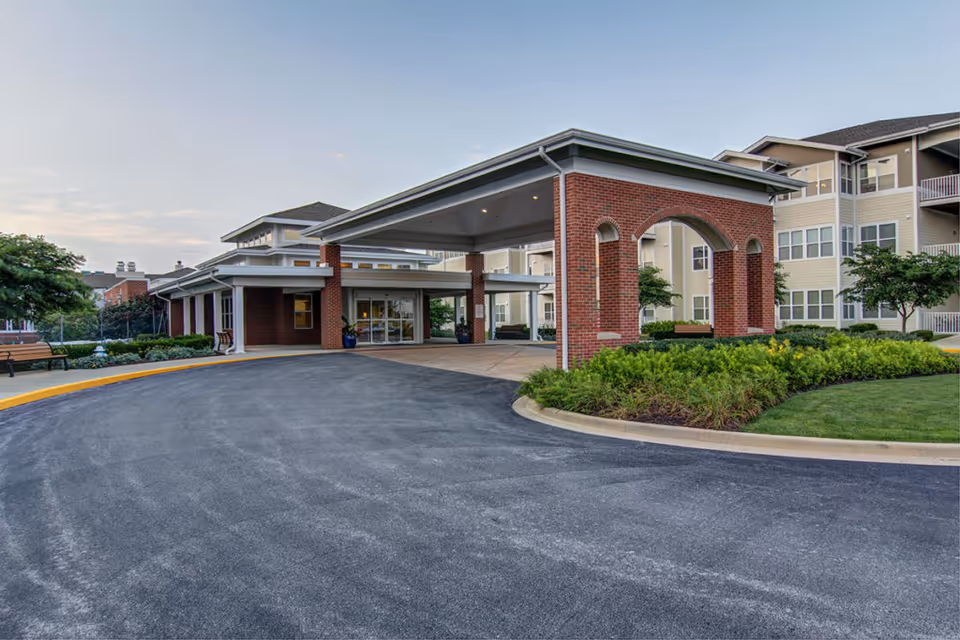 Entrance of a senior living facility with a covered drop-off area supported by brick columns, surrounded by well-maintained landscaping and a paved driveway. The building has multiple stories with numerous windows and balconies.