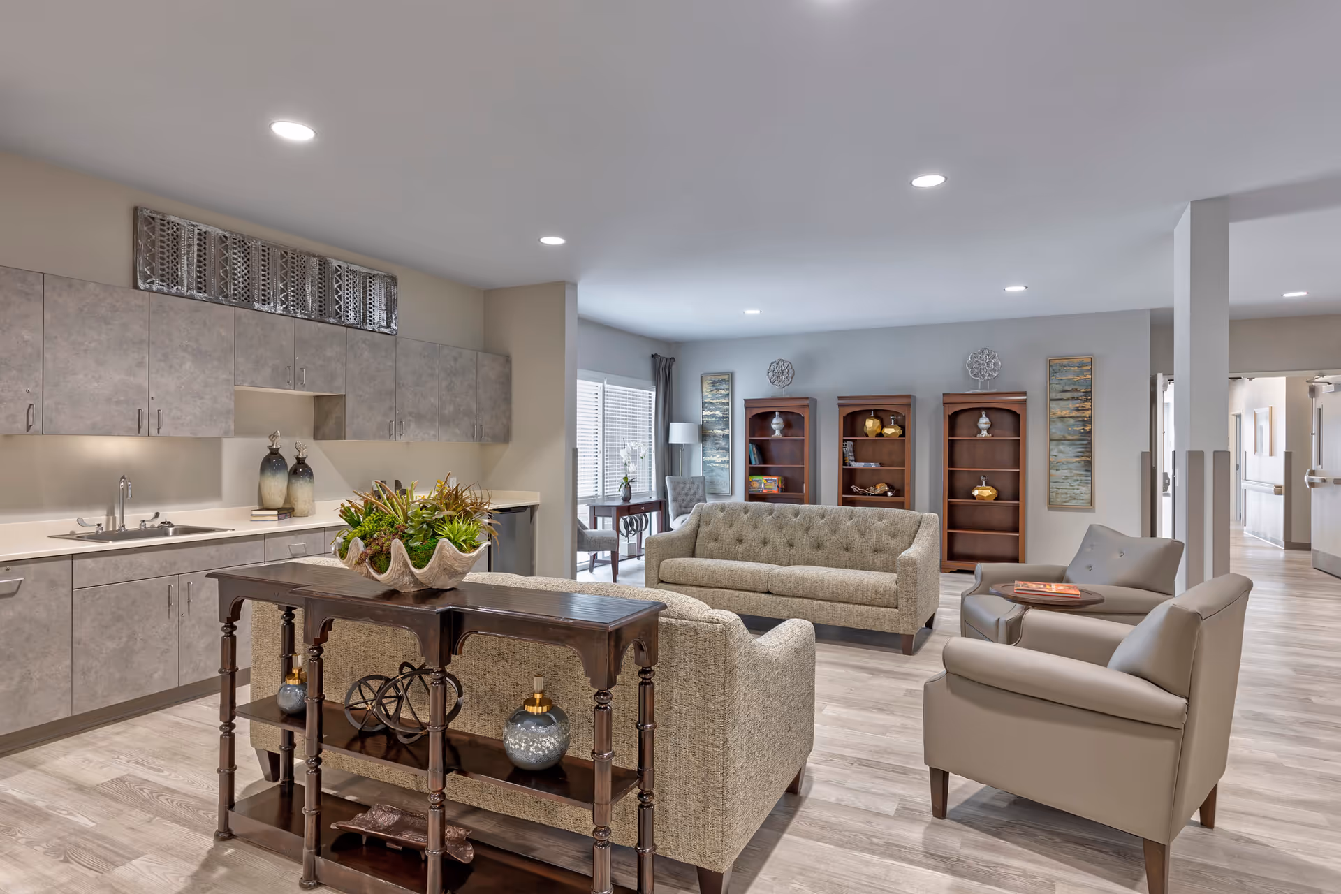 A spacious and well-lit common area in a senior living facility featuring a kitchenette with gray cabinets and a sink on the left, a wooden console table with decorative items, two beige fabric sofas, two gray leather armchairs, and three wooden bookshelves with decorative objects against a light gray wall. The room has light wood flooring and recessed ceiling lights.
