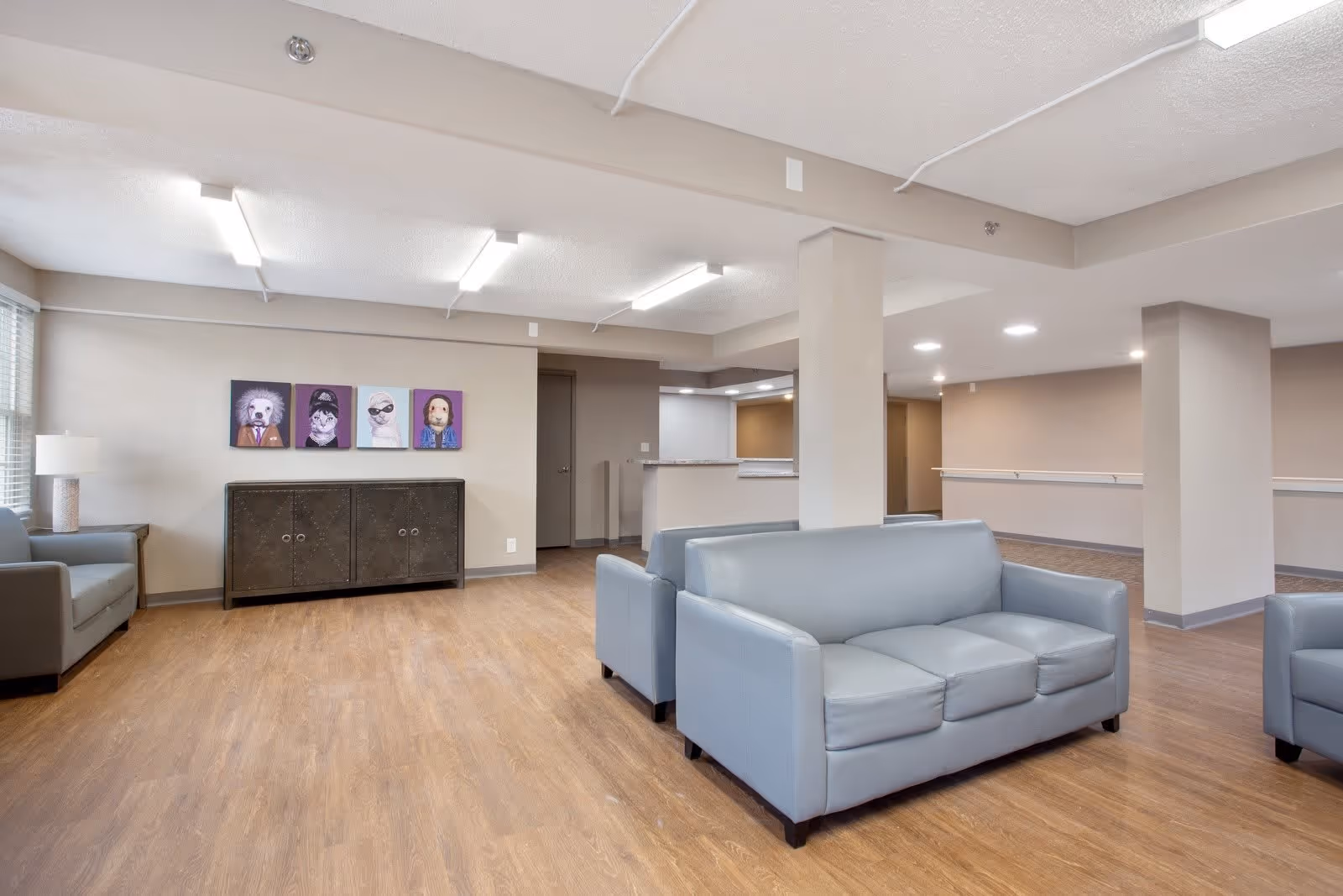A spacious common area with light gray sofas and armchairs on a wood floor. There is a dark cabinet against the wall with four colorful animal portraits above it. The room has beige walls, fluorescent ceiling lights, and a countertop area in the background.