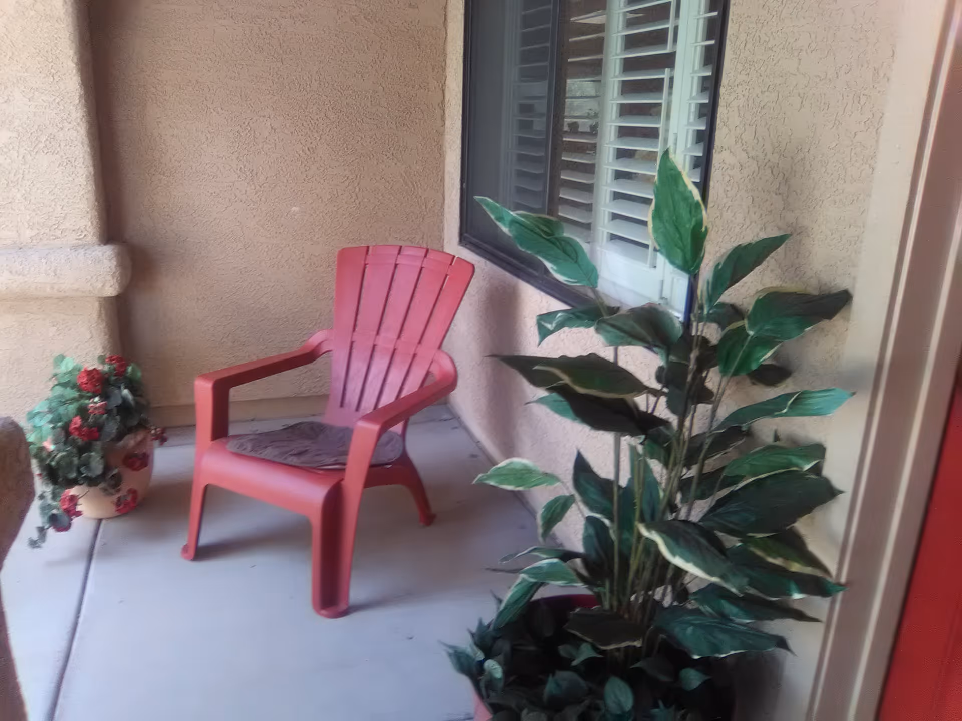 A small outdoor patio area with a red plastic chair that has a cushion on the seat. There are two potted plants, one with red flowers on the left and a larger green leafy plant on the right. The patio has beige stucco walls and a window with white shutters.