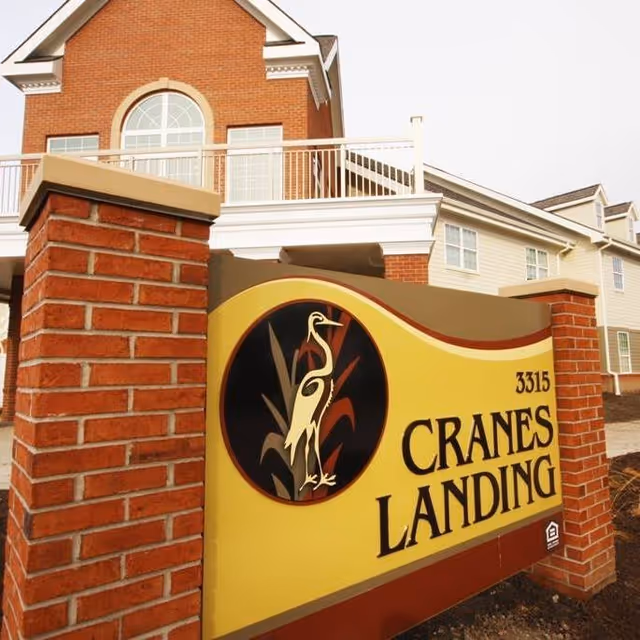 Brick entrance sign reading "Cranes Landing" with a crane logo in front of a multi-story residential building.