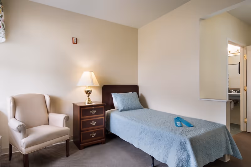A bedroom in a senior facility featuring a single bed, chair, nightstand, and lamp.