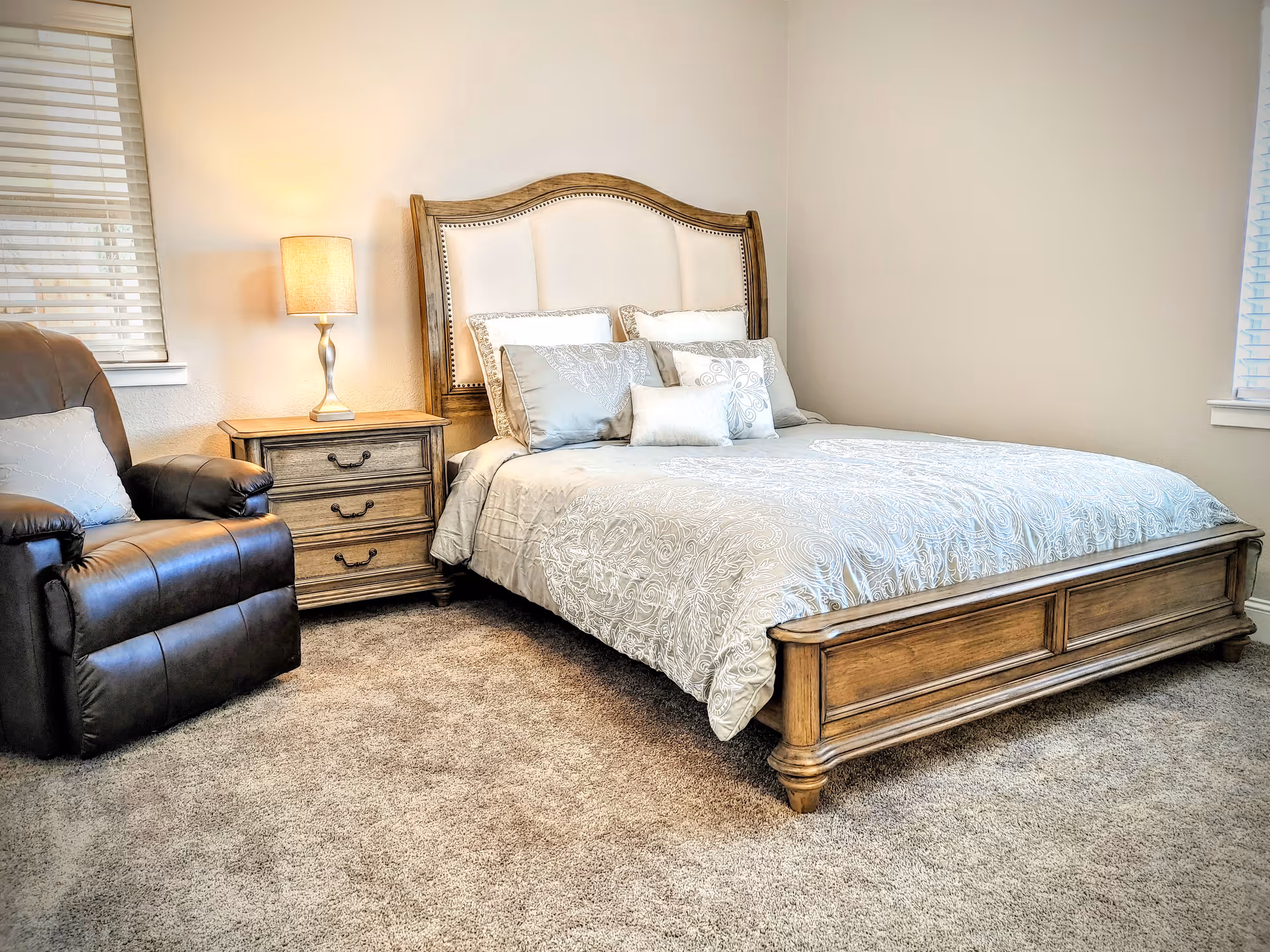 A cozy bedroom with a wooden bed frame and a white and gray patterned comforter. The bed is adorned with multiple pillows. Next to the bed is a wooden nightstand with three drawers and a lamp on top. To the left of the nightstand is a dark brown leather recliner chair. The room has beige walls, carpeted floor, and two windows with blinds.