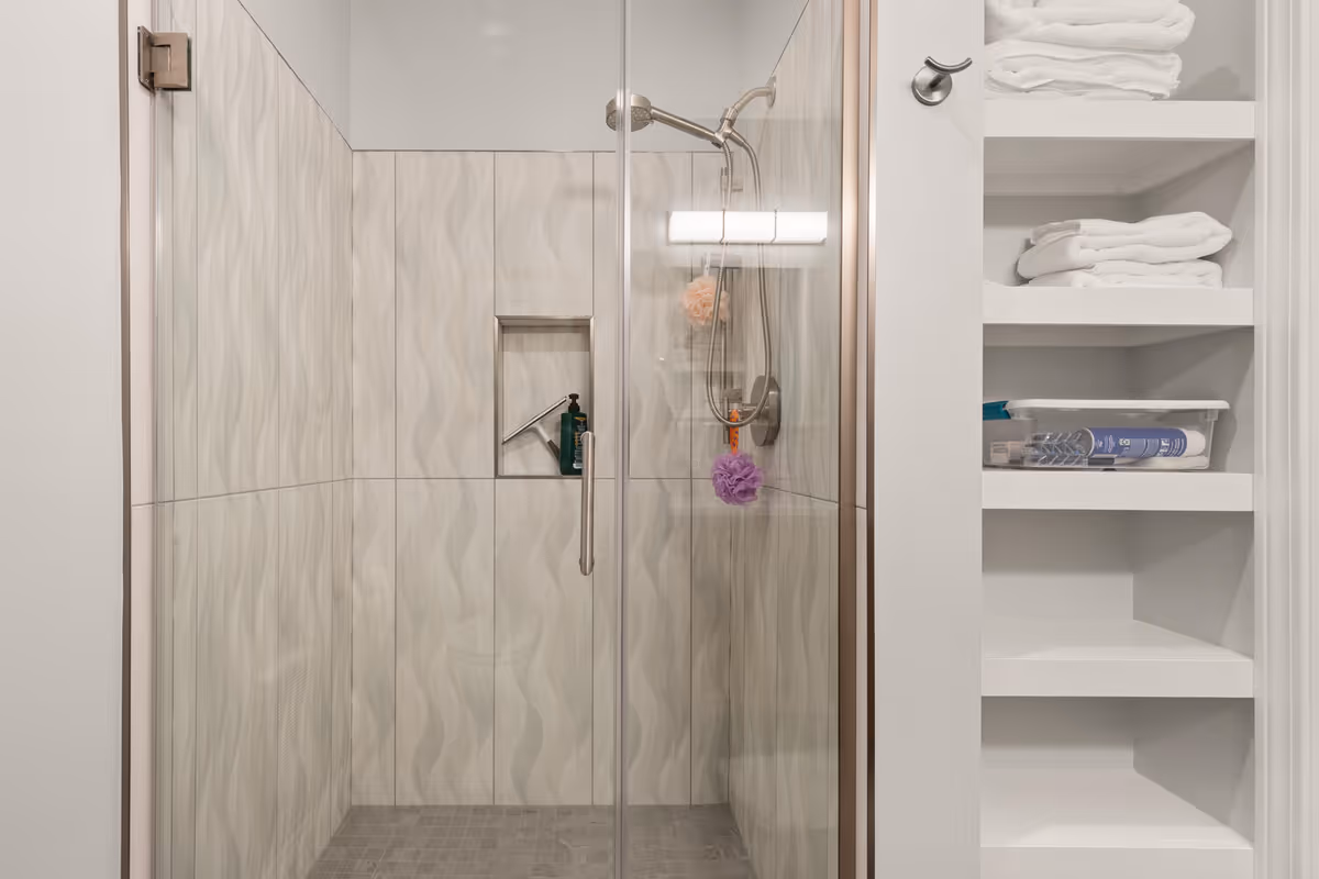 Glass-enclosed walk-in shower with patterned tile walls, a built-in niche and showerhead, next to open shelving stocked with towels.