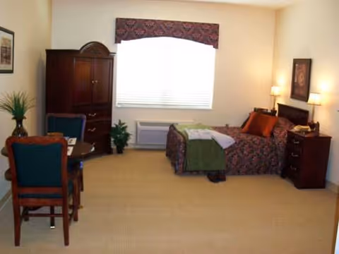 Furnished single-occupancy bedroom with a bed, nightstand, armoire, small table and chairs, and a window with blinds.