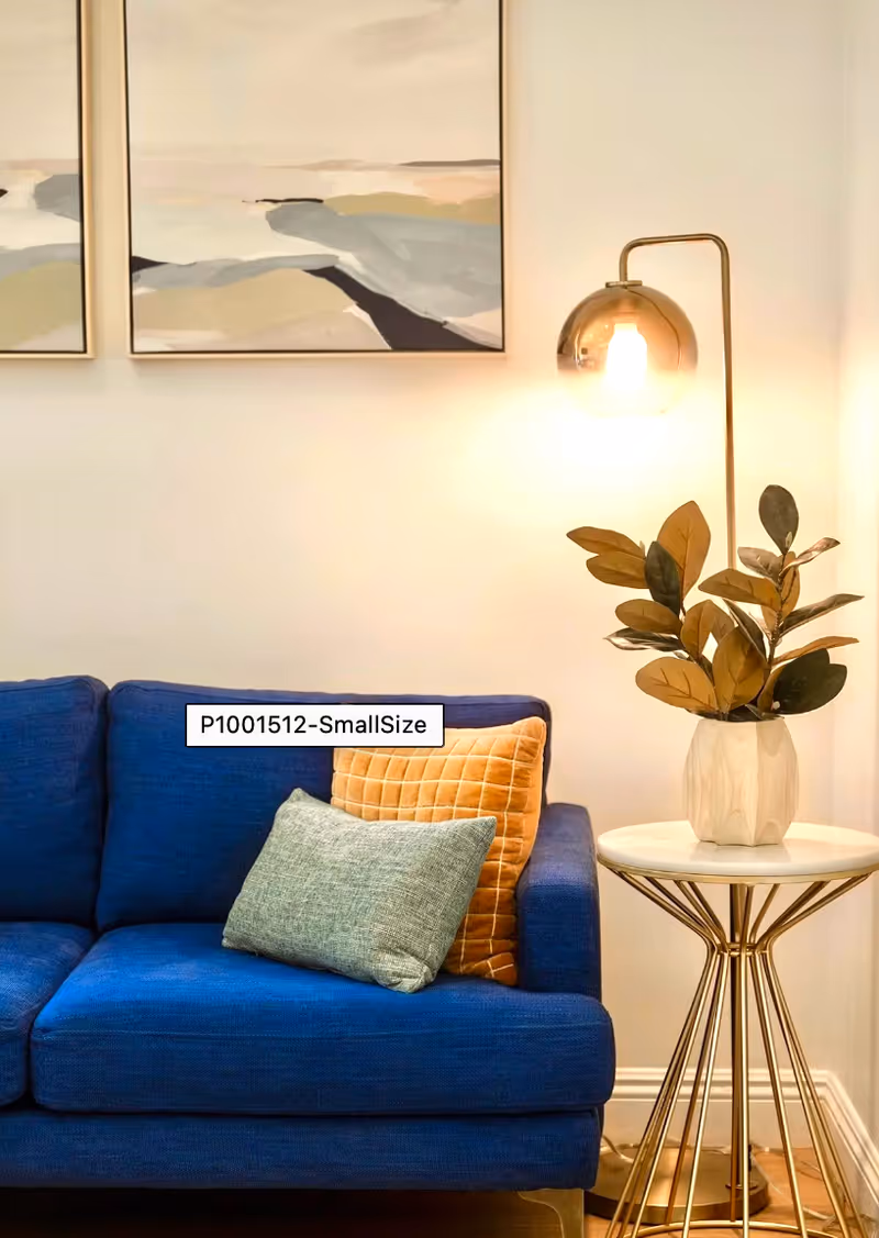 A cozy corner of a living room featuring a blue sofa with two cushions, one orange and one gray. Next to the sofa is a round side table with a white marble top and gold legs, holding a potted plant with green and brown leaves. A modern floor lamp with a gold base and a glowing bulb inside a glass shade stands behind the table. Two abstract landscape paintings hang on the wall above the sofa.