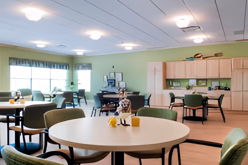 Bright communal dining and activity room with round tables, green upholstered chairs, large windows, and a kitchenette against a green wall.
