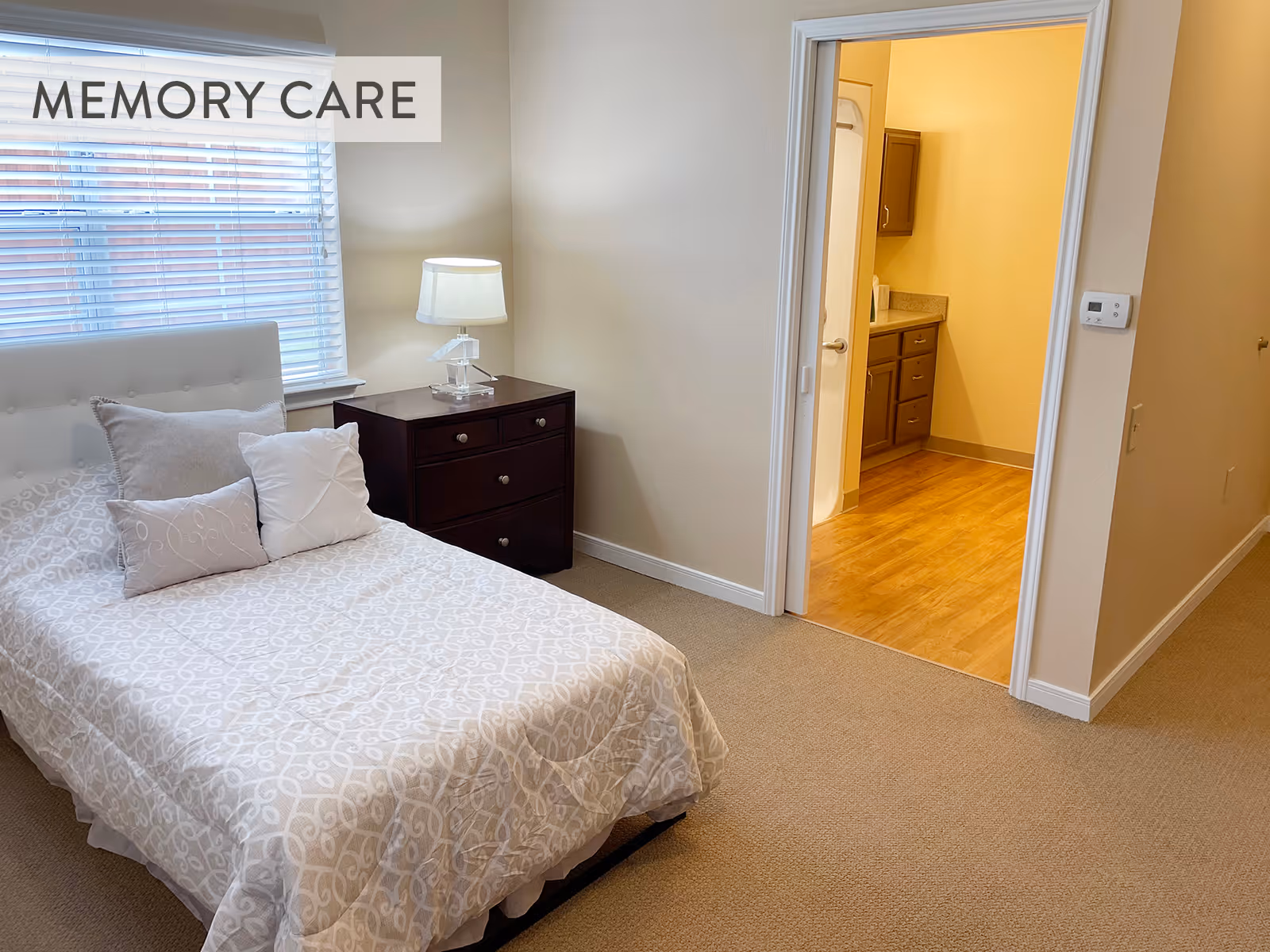 Tidy bedroom with a single bed, nightstand and lamp, and an open doorway leading to a bathroom/vanity area.