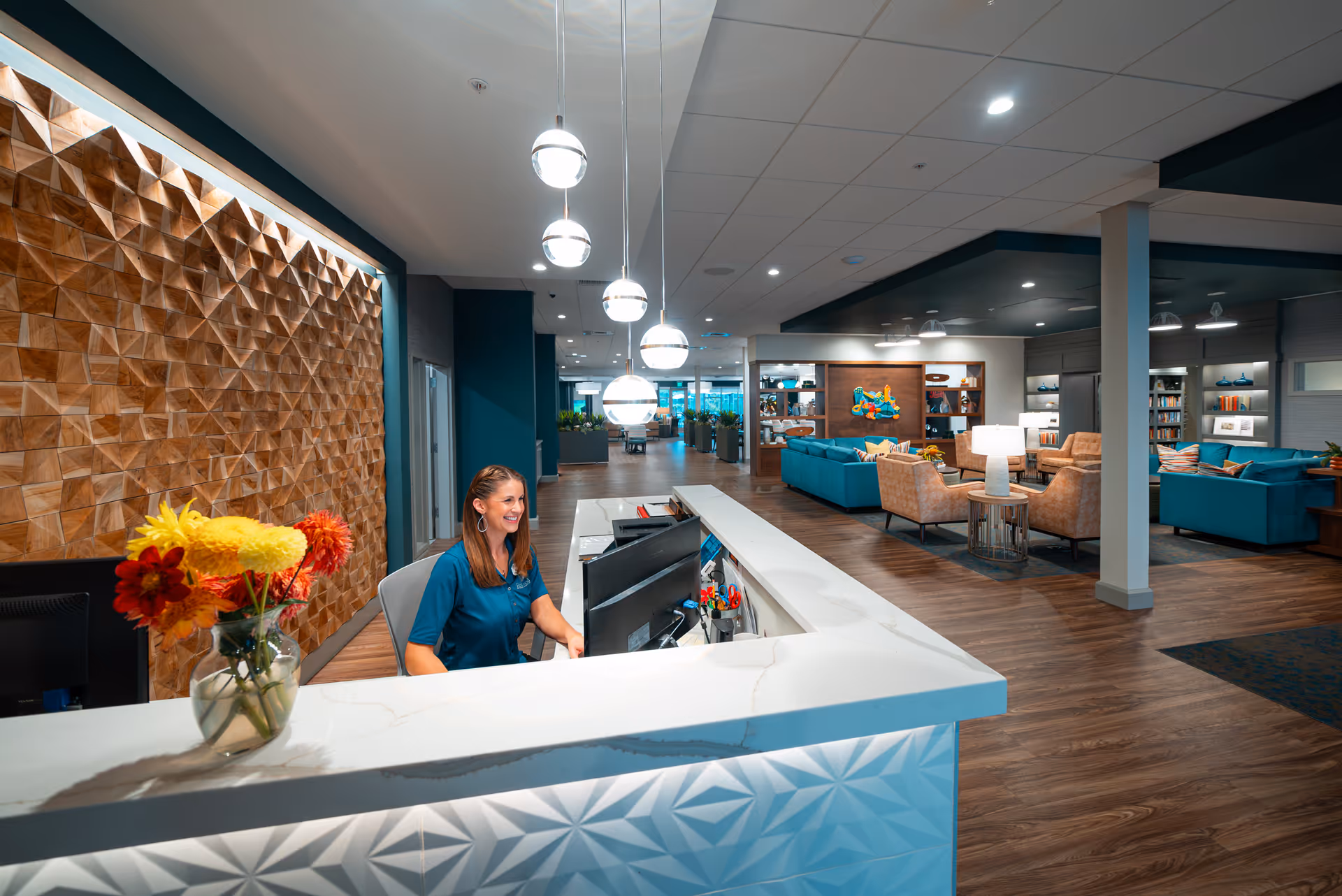 A modern reception area in a senior living facility with a smiling receptionist seated behind a white marble counter. The background features a textured wooden wall panel, hanging spherical lights, and a spacious lounge area with teal sofas, beige armchairs, and bookshelves. The floor is wooden, and the space is well-lit with ceiling lights.