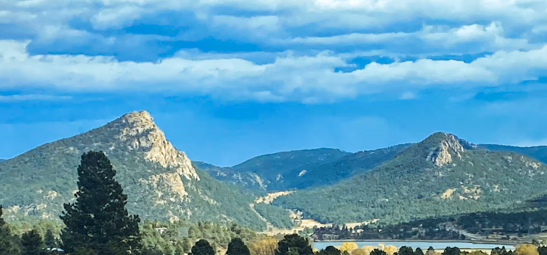 Scenic view of a mountainous landscape with two prominent rocky peaks, green forested hills, and a partly cloudy blue sky above.