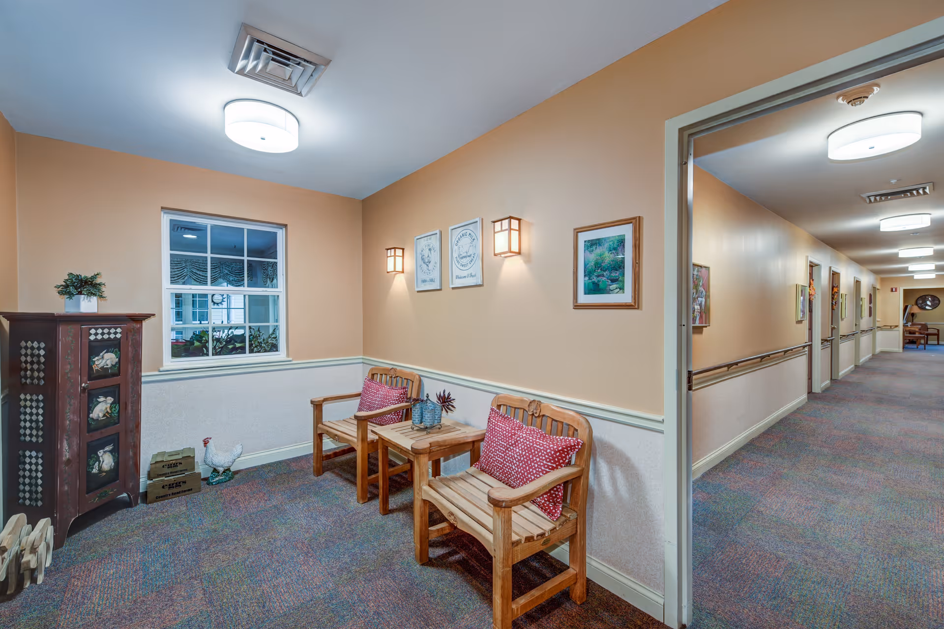 A cozy seating area in a hallway of a senior living facility with two wooden chairs featuring red patterned cushions and a small wooden table between them. The walls are painted beige with framed artwork and wall sconces providing warm lighting. A window is visible on the left side, and the hallway extends to the right with more framed pictures and ceiling lights.