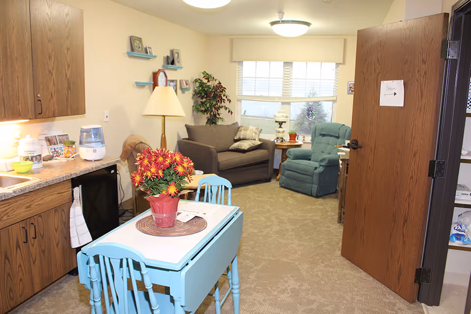 Cozy senior living room with a kitchenette on the left, a small blue dining table with flowers in the foreground, and a sofa and recliner by a window.