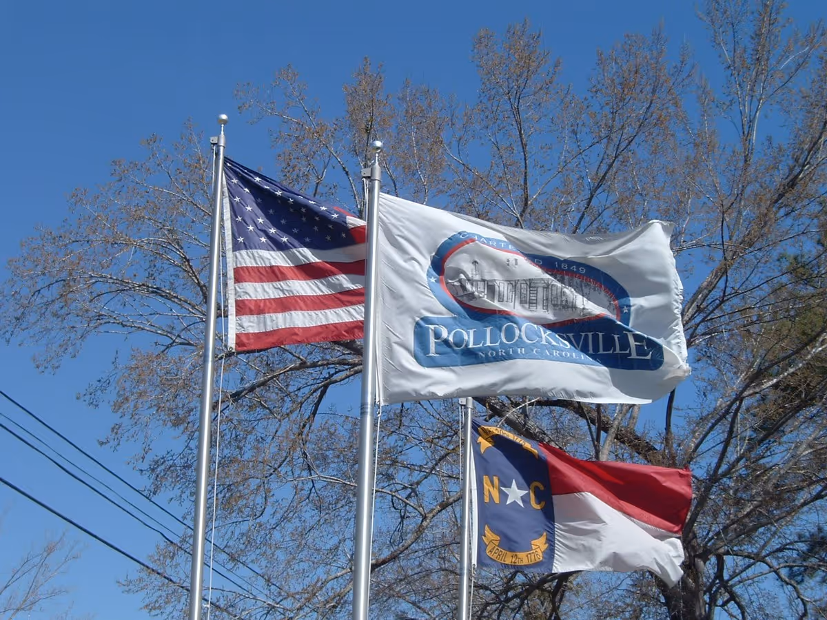 Three flags flying on flagpoles against a clear blue sky with leafless trees in the background. The flags include the United States flag, the Pollocksville North Carolina flag, and the North Carolina state flag.