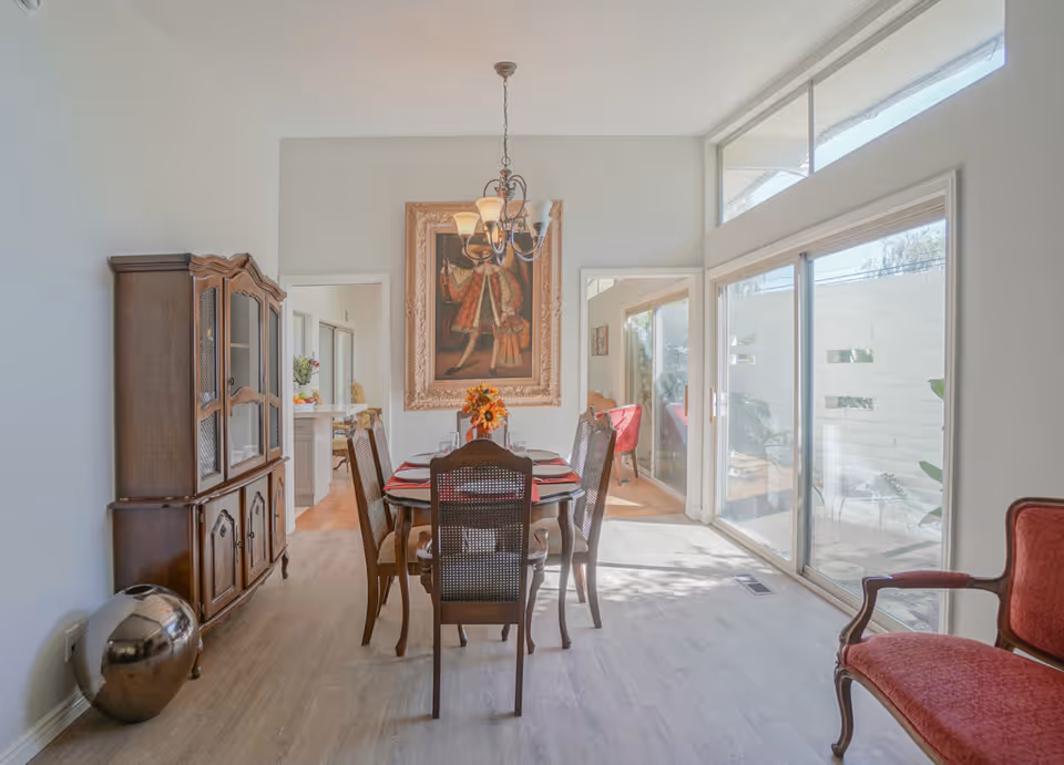 Bright dining room with a wooden table and chairs, a china cabinet, chandelier, large framed painting, and sliding glass doors to a patio.