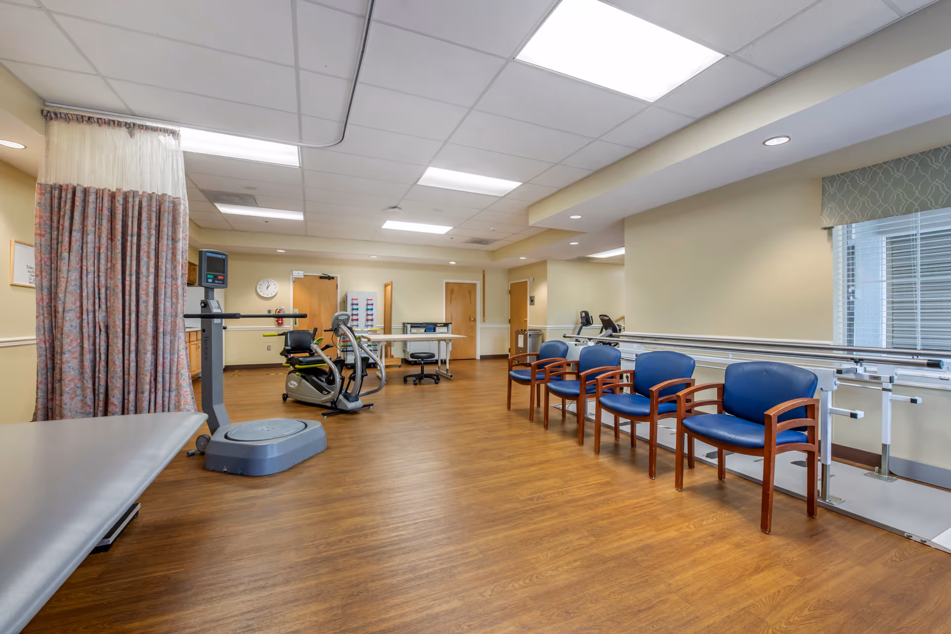 A spacious rehabilitation or physical therapy room with wooden flooring, several blue chairs with wooden armrests lined up against the wall, parallel bars for walking exercises, an exercise bike, a vibration plate machine, a treatment table, and a privacy curtain. The room has beige walls, a window with blinds and a valance, and fluorescent ceiling lights.