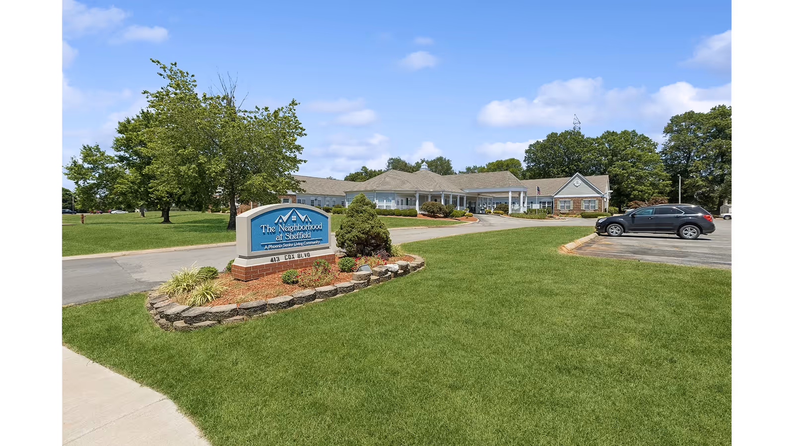 Front exterior of The Neighborhood at Sheffield with a lawn, entrance drive, parking area, and a property sign.