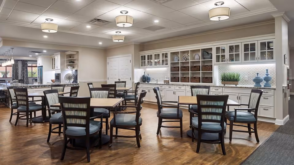 A spacious dining area in a senior living facility with several wooden tables and chairs arranged neatly on a wooden floor. The room features white cabinetry with glass doors displaying decorative items and baskets. The ceiling has recessed lighting and several modern pendant lights. In the background, there is a kitchen area with a large island and additional seating.