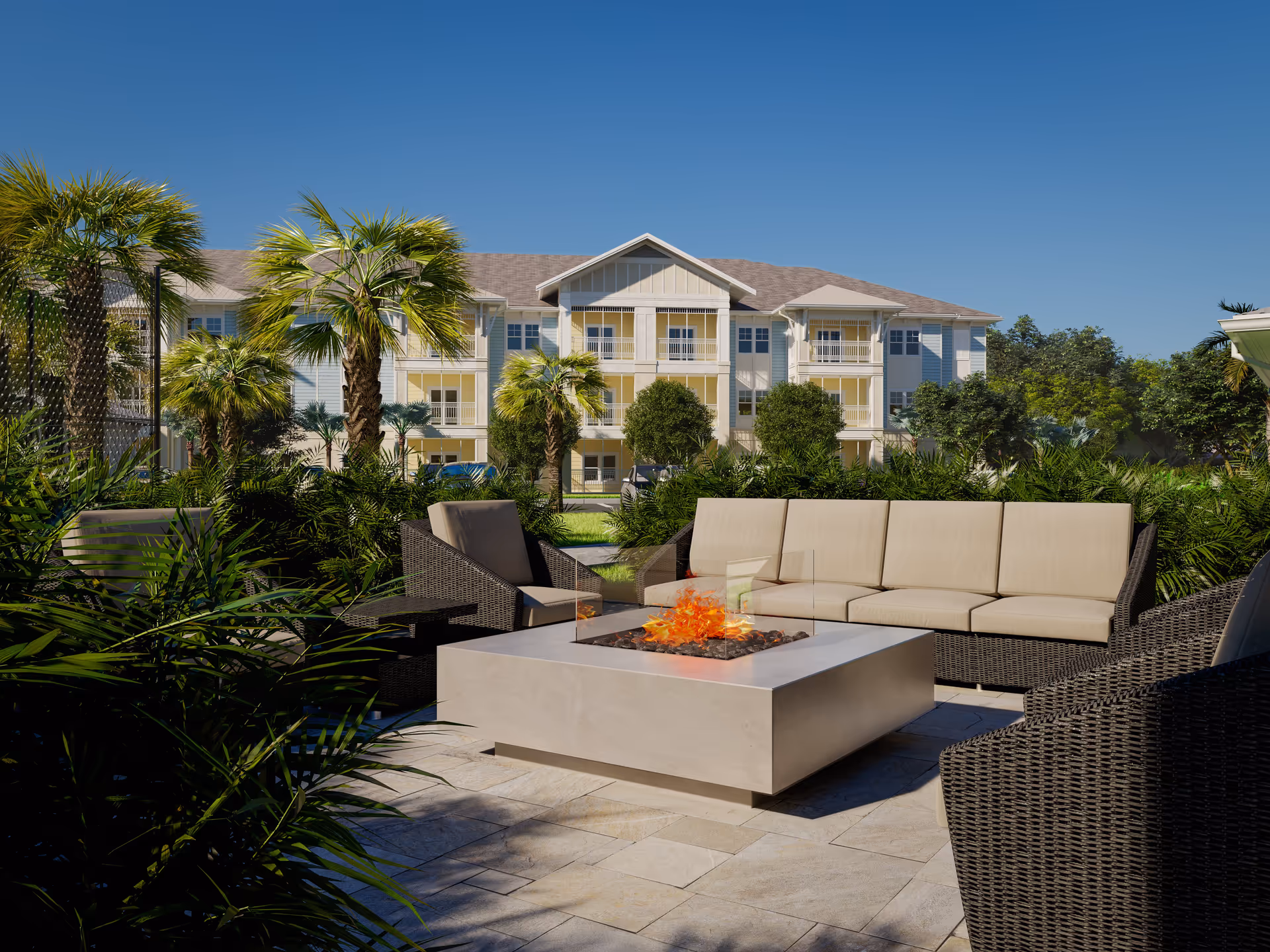 Outdoor seating area with cushioned wicker chairs and a sofa arranged around a modern square fire pit, surrounded by lush greenery and palm trees, with a multi-story residential building in the background under a clear blue sky.
