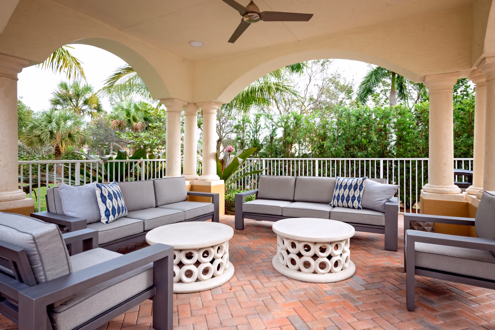 Covered outdoor patio area with cushioned gray sofas and armchairs arranged around two round white tables. The patio has brick flooring, cream-colored columns, and a ceiling fan. Greenery and palm trees are visible beyond the white railing surrounding the patio.
