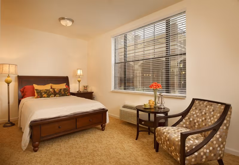 A cozy bedroom with a wooden bed frame, white bedding, and decorative pillows. There are two lamps, one floor lamp and one table lamp on a nightstand. A window with blinds lets in natural light. Next to the window is a small round table with a vase of red flowers and a patterned armchair.