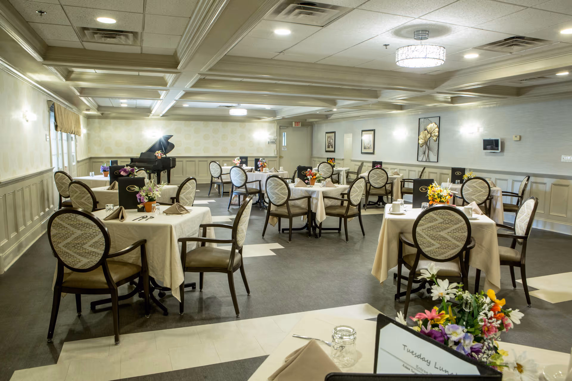 Dining room with multiple set tables and chairs, floral centerpieces, and a grand piano in the background.
