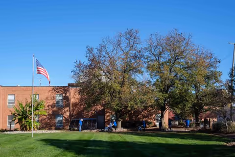 Exterior view of a brick building with several windows, a green lawn in front, and an American flag on a flagpole. There are large trees with autumn foliage and a few people walking or sitting near the building.