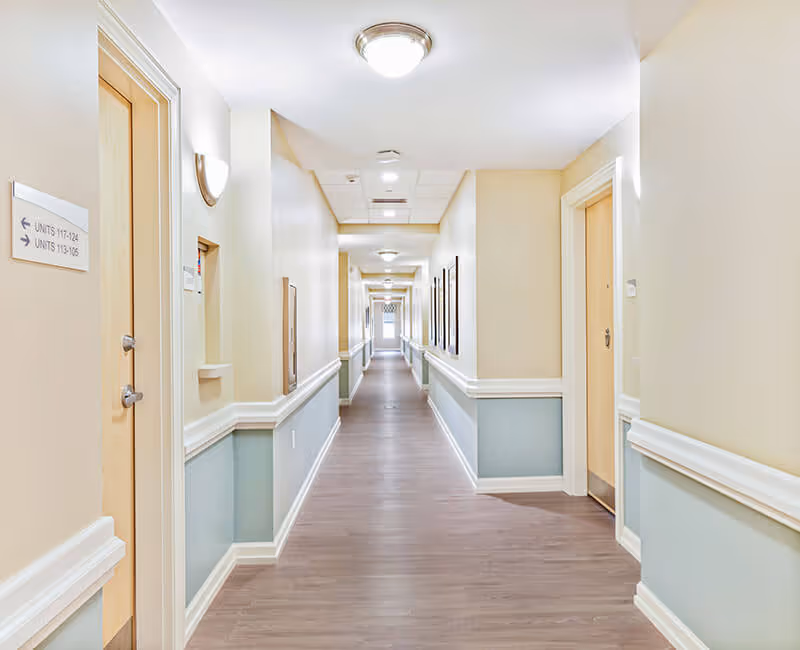 A long, well-lit hallway in a senior living facility with light beige walls and light blue wainscoting. Several doors line the corridor, and there are signs indicating unit numbers. The floor is covered with wood-like flooring, and ceiling lights illuminate the space evenly.