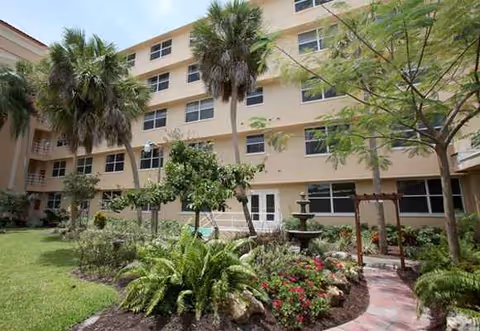 Three-story beige senior living building with palm trees, a garden, and a decorative fountain courtyard.