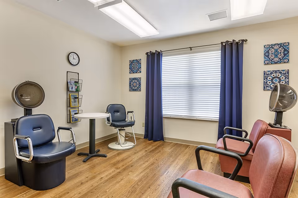 A small salon room with two black salon chairs with hair dryers, two red chairs, a small round table, a wall clock, and a window with blue curtains and white blinds. The floor is wooden and there are decorative blue patterned wall hangings.