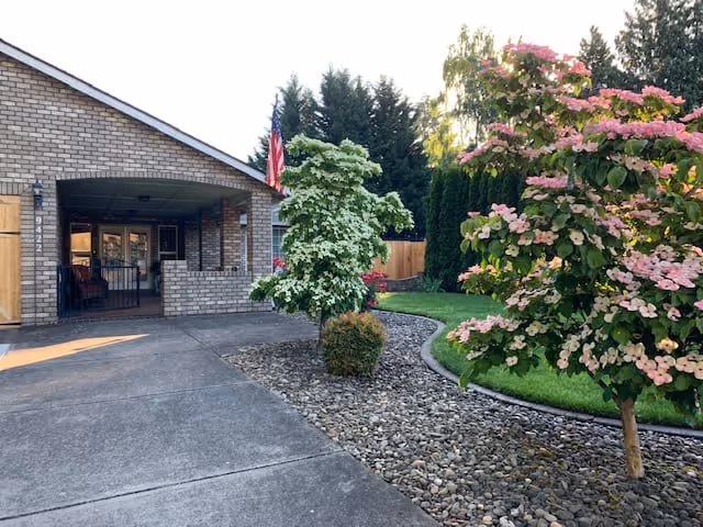 Exterior view of a brick building with a covered entrance, a concrete driveway, and landscaped garden area featuring small trees with pink and white flowers and green grass.