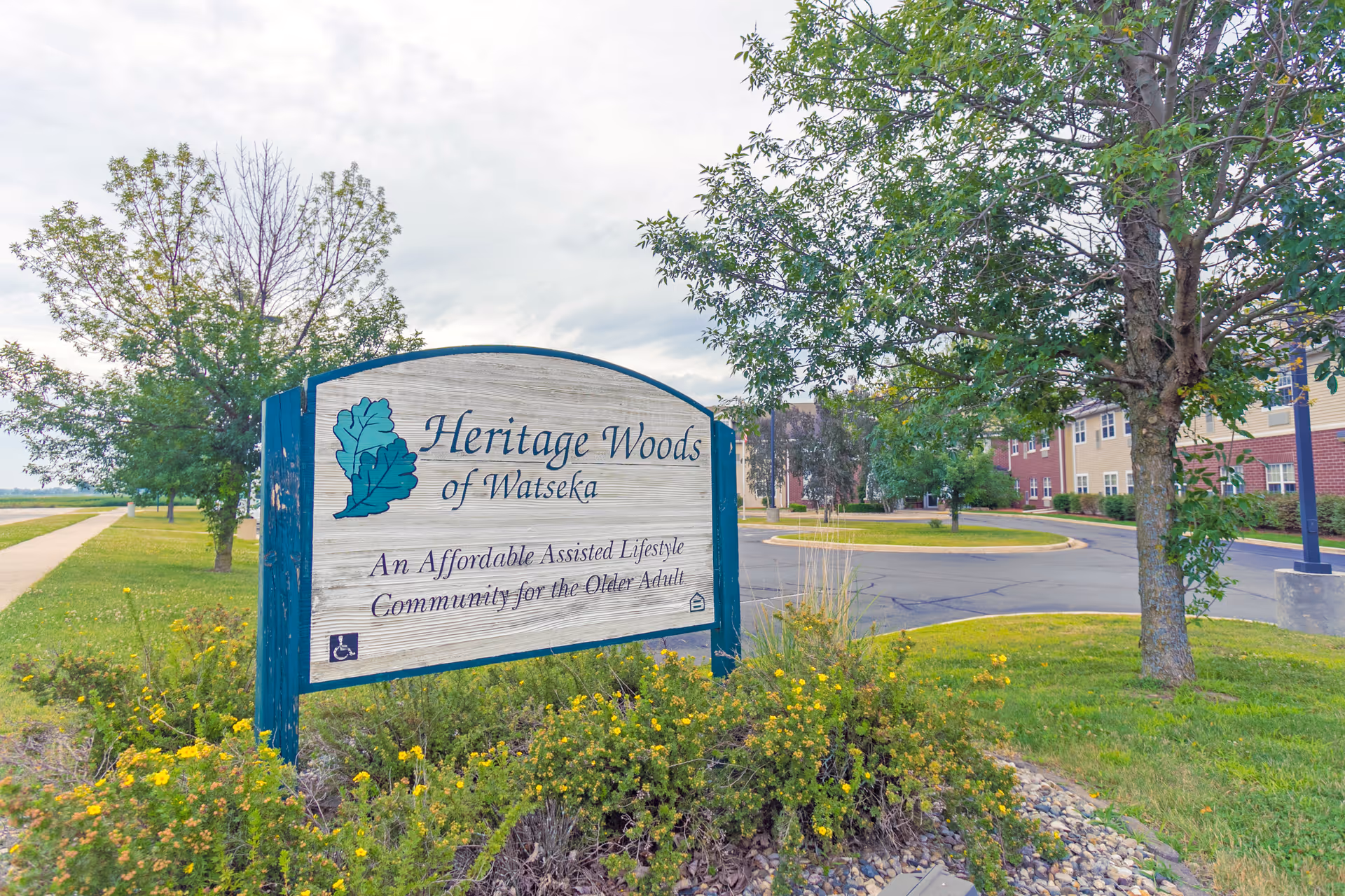 Entrance sign for Heritage Woods of Watseka on landscaped grounds with trees and the facility building and driveway in the background.