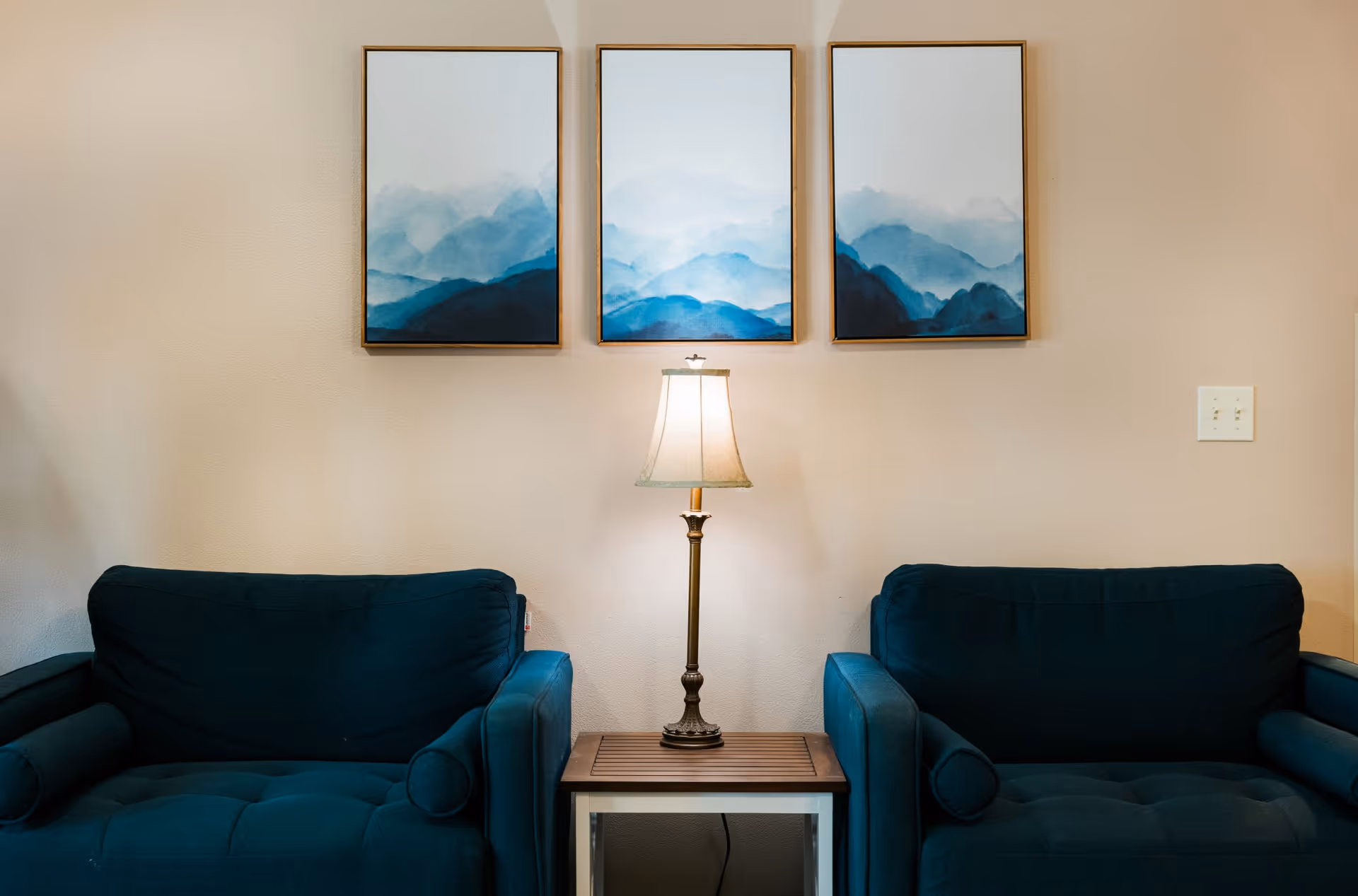 Two dark blue armchairs separated by a small wooden side table with a lamp on it. Above the lamp, there are three framed paintings depicting abstract blue mountain landscapes on a beige wall.