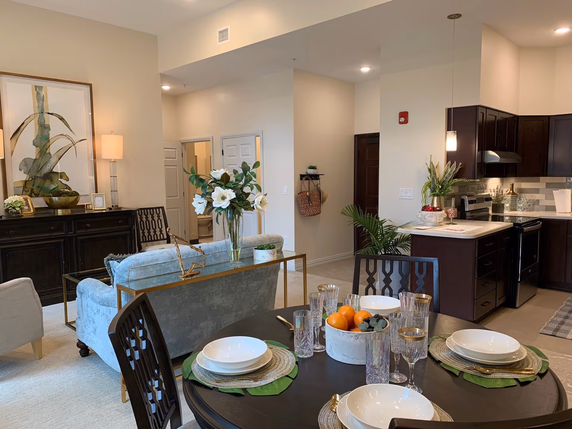 Interior view of a senior living facility showing a dining area with a round table set with plates, glasses, and a centerpiece of fruit. Behind the dining area is a living room with a light blue sofa, a sideboard with a lamp and artwork, and a kitchen with dark wood cabinets and stainless steel appliances. The space is well-lit with ceiling lights and decorated with plants and flowers.