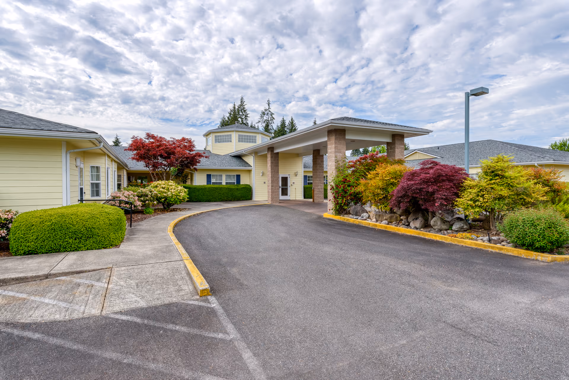 Exterior view of Easthaven Villa Assisted Living & Memory Care building with a covered entrance, surrounded by well-maintained landscaping including bushes and colorful trees under a partly cloudy sky.