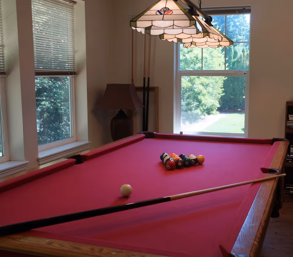 Sunlit interior room with a red-felt pool table, racked billiard balls, a cue stick, windows, and a hanging lamp.
