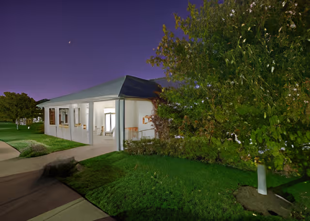 Exterior view of a single-story building at night with a gray roof and white walls, surrounded by green grass and trees under a dark purple sky.