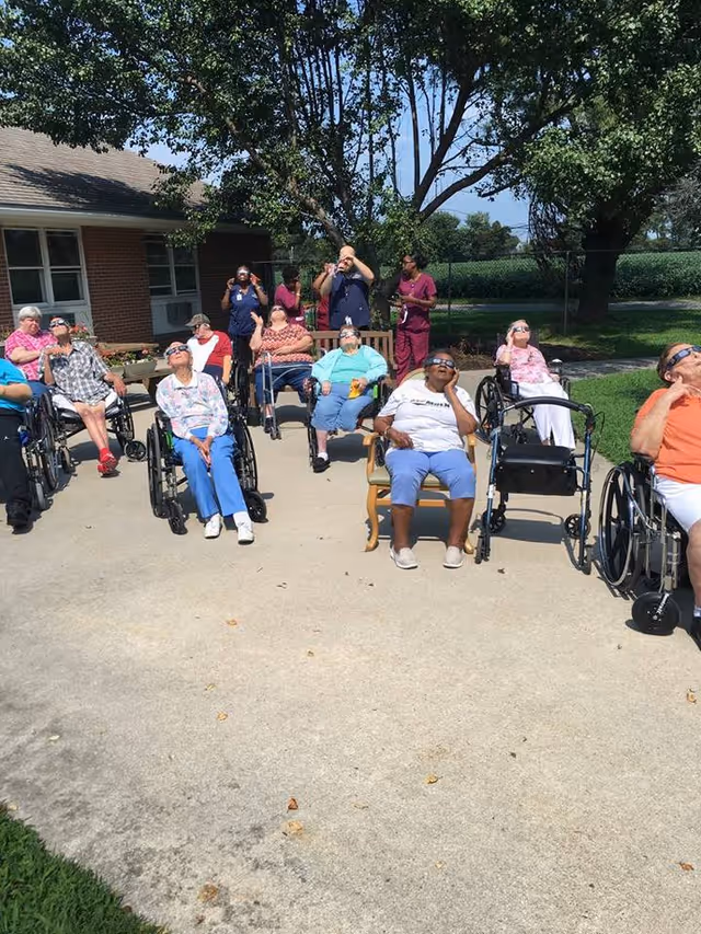 A group of elderly individuals, many in wheelchairs, sitting outside on a paved area under a tree, all wearing protective glasses and looking up at the sky. Several caregivers stand behind them, also looking up. The setting appears to be a sunny day at a senior living facility with a brick building and green fields in the background.