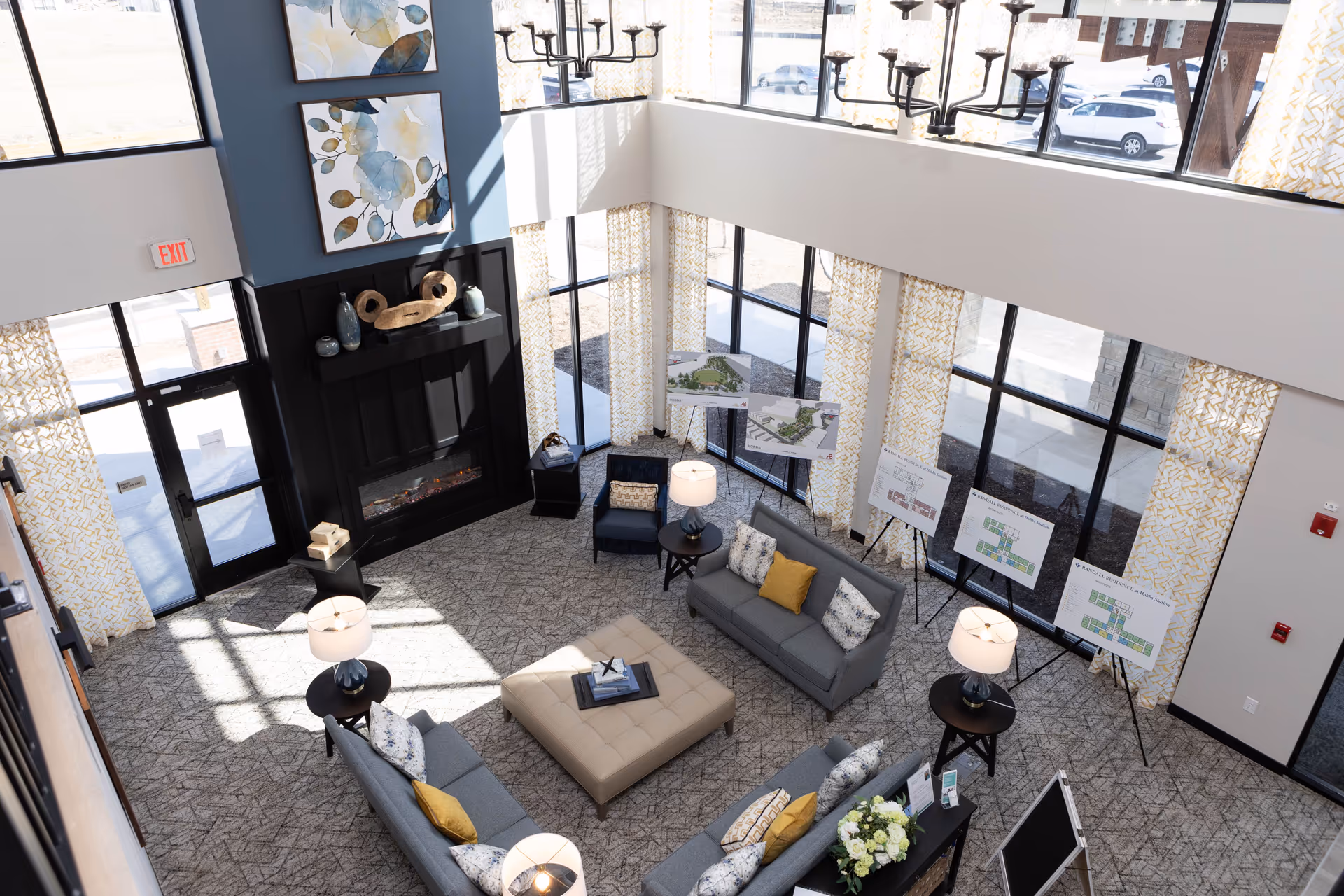 A bright and spacious senior living facility common area with large windows, patterned yellow and white curtains, and a high ceiling. The room features a cozy seating arrangement with gray sofas, a beige ottoman, side tables with lamps, and decorative pillows. There is a modern black fireplace with decorative items on the mantel and two framed floral artworks above it. Several easels display floor plans and facility layouts near the windows.
